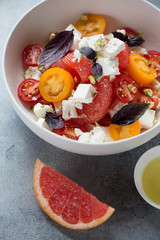 Close-up of salad with grapefruit, feta cheese, red and yellow cherry tomatoes, nuts and red basil leaves served in a white bowl, studio shot