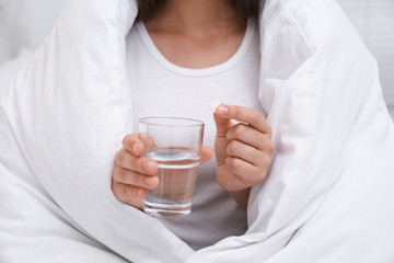 Woman holding glass of water and pill in bedroom, closeup