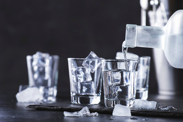 Vodka in shot glasses pouring out of the bottle on black stone background, iced strong drink in misted glass