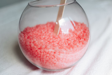 Closeup of a round glass vase with pink wax granules for depilation and wooden stick on a white background.