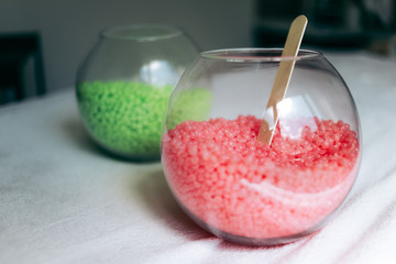 Two round glass vases with pink and green wax granules for depilation and wooden stick in a treatment room.