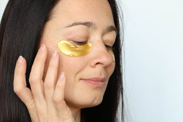 face of middle aged woman with fingers applying golden collagen patches under eyes, on whitebackground. Beauty treatment