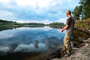 Fototapeta premium Caucasian adult man fishing in evening with beautiful reflection sky and clouds on the river.
