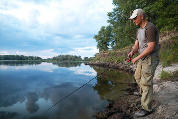 Caucasian adult man fishing in evening with beautiful reflection sky and clouds on the river.