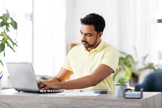 remote job, technology and people concept - young indian man with laptop, calculator and papers working at home office