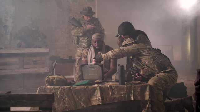 Detachment of military men in camouflage discussing plan of attack and using army laptop. Group of professional soldiers based in abandoned building full of smoke planning assault on enemy camp