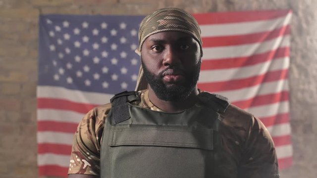 Portrait of serious african american military male in camouflage bandana and bulletproof vest posing against of USA flag. Brave bearded army male standing on background of US flag
