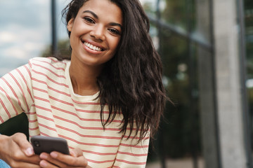 Image of smiling african american girl using mobile phone while sitting