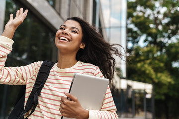 Image of african girl waving hand and holding laptop while walking