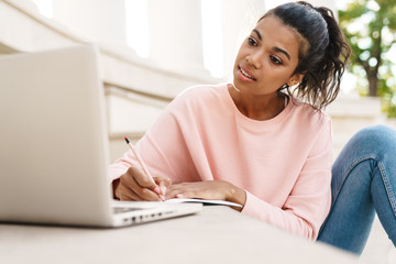 Image of pleased african american student girl doing homework with laptop