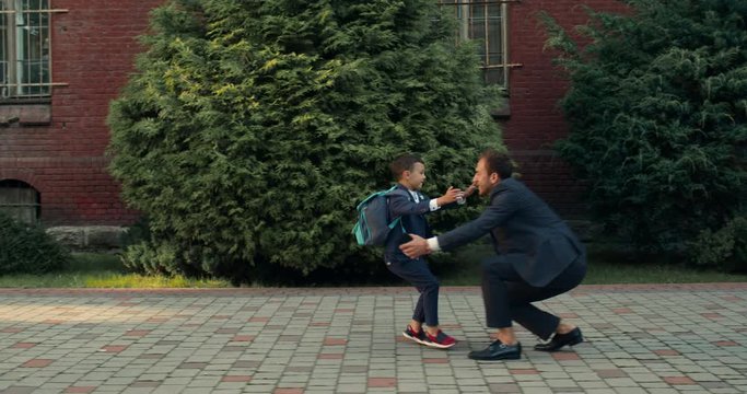 Side view of teen boy in school uniform running into dad hugs.Cheerful father wearing suit waiting for his son after lessons near school building. Concept of happy family.