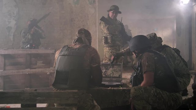Squad of armed military men in camouflage gathering around army laptop displaying local map. Professional soldiers based in abandoned building monitoring enemy forces using map on computer