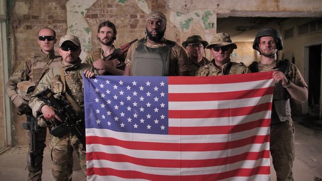 Serious multinational males soldiers in camouflage, bulletproof vests posing with US flag. Brave patriots of USA armed forces standing with weapons and holding symbol of United States of America