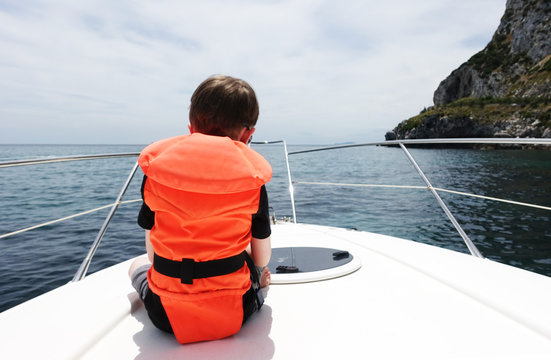 Rear view of boy sitting on front of motor boat wearing orange life jacket
