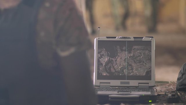 Focus on display of army grade laptop with satellite map of area. Soldiers using special computer to detect enemy forces in collision zone, intelligent military technology concept