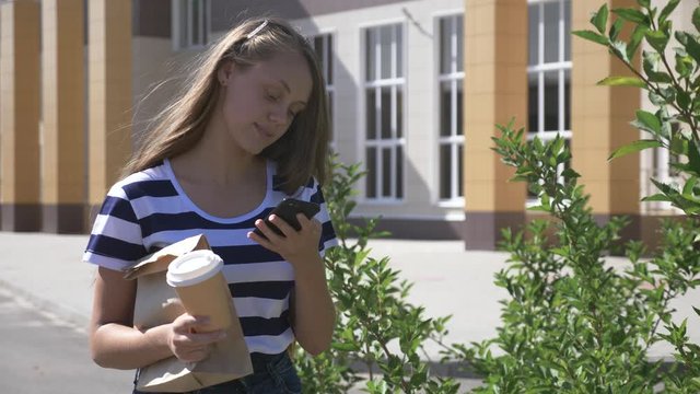 The girl holds a smartphone in her hand. Using smartphone apps to deliver fast food and coffee. Studen walks at lunchtime. A valanter girl helps to deliver food. Social work.