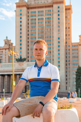 A young blond man in shorts against the background of the main University of Moscow, a high-rise building against the blue sky