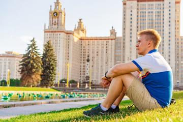 Fototapeta premium A young blond man sits on the grass in front of the main University of Russia, Moscow high-rise