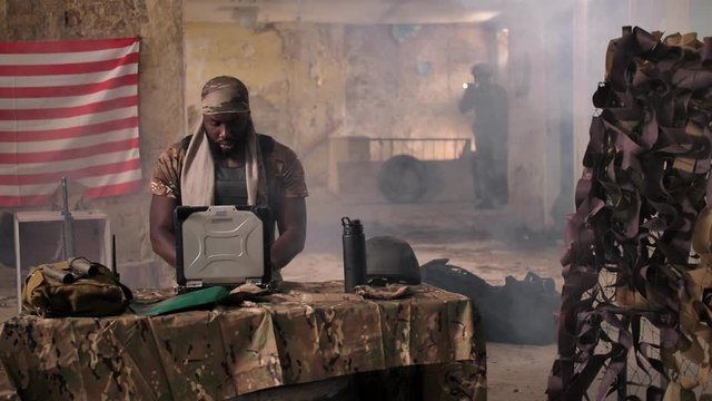 Dark-skinned soldier using army grade laptop for tracking target and walkie-talkie for communication. African american military man working at table in temporary base located in abandoned building