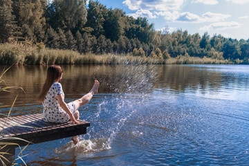 Woman on the lake. Young woman wearing white dress sitting on a wooden pier on the lake and dip her feet in water, creating splashes