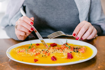 Woman in a grey jumper is eating at the restaurant a beautifully decorated pumpkin dish with red beetroot cubes, greens and seeds. Close-up of woman's hands with red manicure holding cutlery.