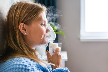 Portrait of a young blonde woman doing inhalation with steam nebulizer at home. Asthma, flu, health care concept.