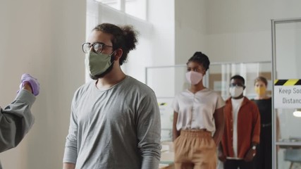 Diverse male and female colleagues in face masks walking in office and keeping social distance while manager scanning their foreheads with digital infrared thermometer during coronavirus pandemic - Powered by Adobe