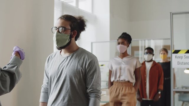 Diverse male and female colleagues in face masks walking in office and keeping social distance while manager scanning their foreheads with digital infrared thermometer during coronavirus pandemic