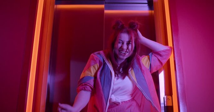 Bottom view of joyful young woman in trendy sports suit looking to camera. Attractive female clubber with stylish hairdo laughing and posing while standing near elevator in neon lights