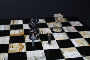 kleine Figuren von Geschäftsleuten auf einem Schachbrett mit Schachfiguren im Hintergrund 