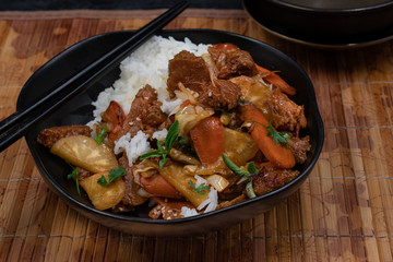 Seitan, daicon and carrot stir fry in black bowl with rice on bamboo mat