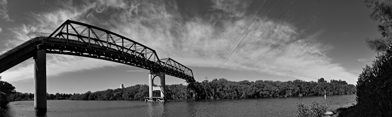 Beautiful black and white panoramic view of a river with tall pedestrian and water pipe bridge, and rainbow look-alike clouds, Parramatta river, Rydalmere, Sydney, New South Wales, Australia