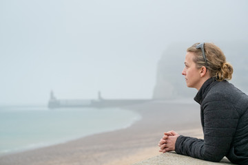 Woman wearing warm clothes and a purse looking out over a foggy sea from a pebble beach on the shore