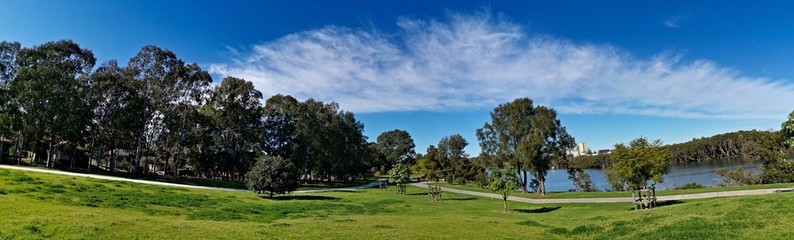 Fototapeta na wymiar Beautiful panoramic view of a park with green grass, tall trees, walking trail and rainbow look-alike clouds, Reid Park, Parramatta Cycleway, Rydalmere, Sydney, New South Wales, Australia 