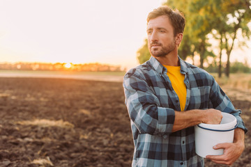 Naklejka na ściany i meble rancher in plaid shirt holding bucket while standing on plowed field in sunshine