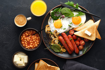 Traditional full English breakfast with fried eggs, sausages, beans, mushrooms, grilled tomatoes on black background.