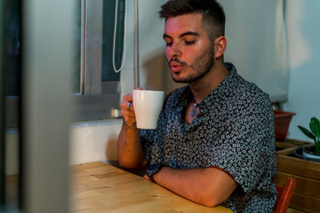 Joven con taza de cafe trabajando en el ordenador junto a la ventana