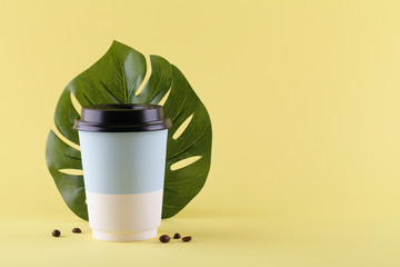 take away paper cup for coffee with coffee beans and monstera leaf on yellow background. copy space. minimal coffee concept.