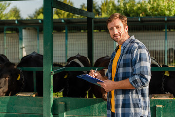 Fototapeta na wymiar farmer in plaid shirt looking at camera while standing near cowshed and writing on clipboard