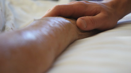 Grandson holds and comforts hand of his old grandmother in medical clinic. Young man gently touches wrinkled arm of sick mature grandma lying in bed hospital. Son shows care and love to his mother