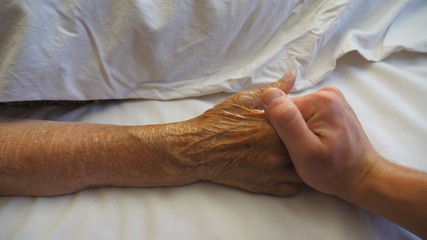 Young man funny takes and comforts hand of his elderly grandmother in hospital. Grandson gently touches wrinkled arm of sick mature woman in medical clinic. Son gives support to his mother. Top view