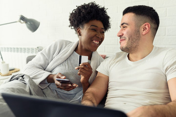 Happy young couple is doing online shopping, lying in a bed, using credit card and laptop