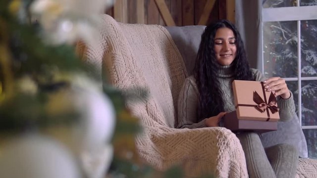 Family christmas, Fun party, Stay at home, New Year celebration. A woman opens a gift box and rejoices sitting in a chair at home on Christmas Eve