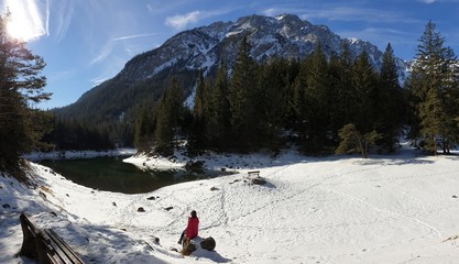A woman sitting at the bench next to the shore of Green Lake in Austrian Alps. Mountains and ground are covered with snow. Winter hiking. She is enjoying a frosty winter day. Winter wonderland
