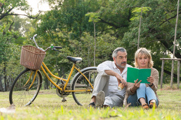 Happy senior caucasian couple man and woman sitting on grass reading book at park with bicycle background, Happy retirement elderly spending and relaxing time together concept, banner copy space