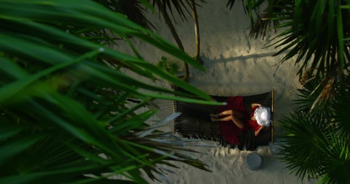 Beautiful young woman lounges and relaxes swaying on hammock surrounded by palm trees