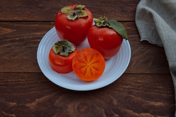 Red, ripe, juicy and soft persimmon on a white plate whole and cut in half with green leaves on a brown wooden background with a linen napkin, seasonal fruit concept, autumn harvest, organic food and 