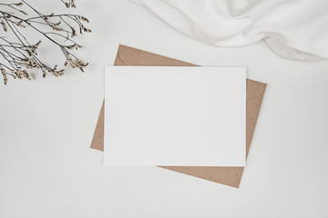 Blank white paper on brown paper envelope with Limonium dry flower and white cloth. Mock-up of horizontal blank greeting card. Top view of Craft envelope on white background. Flat lay minimalism