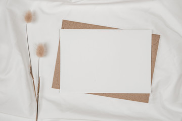 Fototapeta premium Blank white paper on brown paper envelope with Rabbit tail dry flower on white cloth. Mock-up of horizontal blank greeting card. Top view of Craft envelope on white background. Flat lay minimalism