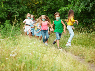 Fototapeta premium Happy little kids wearing protective face mask jumping and running on meadow, forest. Looks happy, cheerful, sincere. Copyspace. Childhood, pandemic concept. Healthcare, coronavirus pandemic.